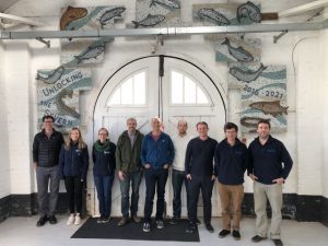 9 members of the Unlocking the Severn team and scientific monitoring group stand in front of the double doors of the workshop. Behind them, the mosaic of fish made by Cherry Orchard primary school frames the doorway.